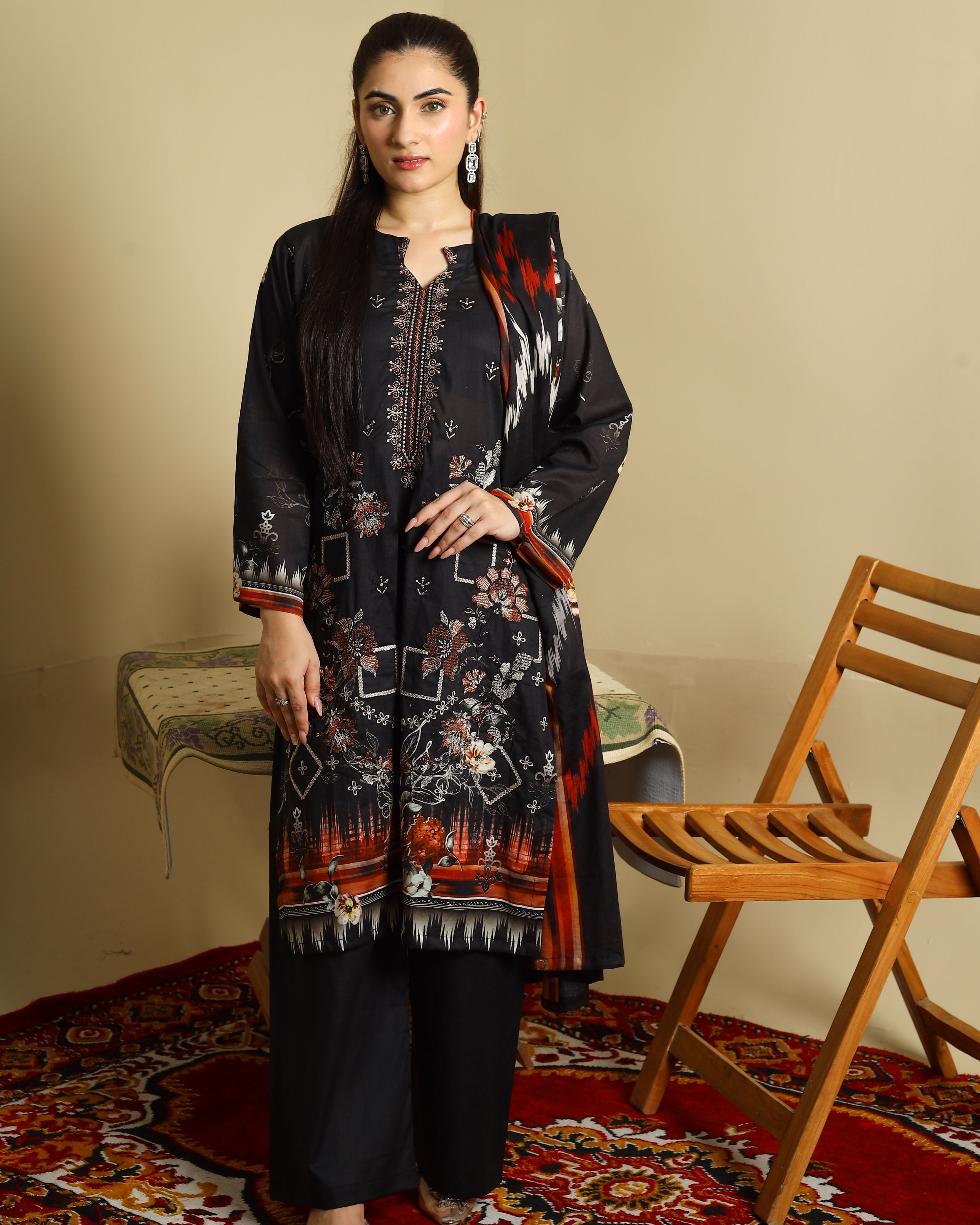 A woman models a three-piece black ethnic suit set featuring a printed kurta, wide-leg trousers, and a matching dupatta, styled in a room with a wooden chair and patterned rug.