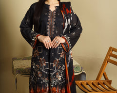 A woman modeling a black three-piece ethnic suit with intricate red, orange, and white floral and geometric prints, standing in a room with a beige wall, a wooden chair, and a small table.